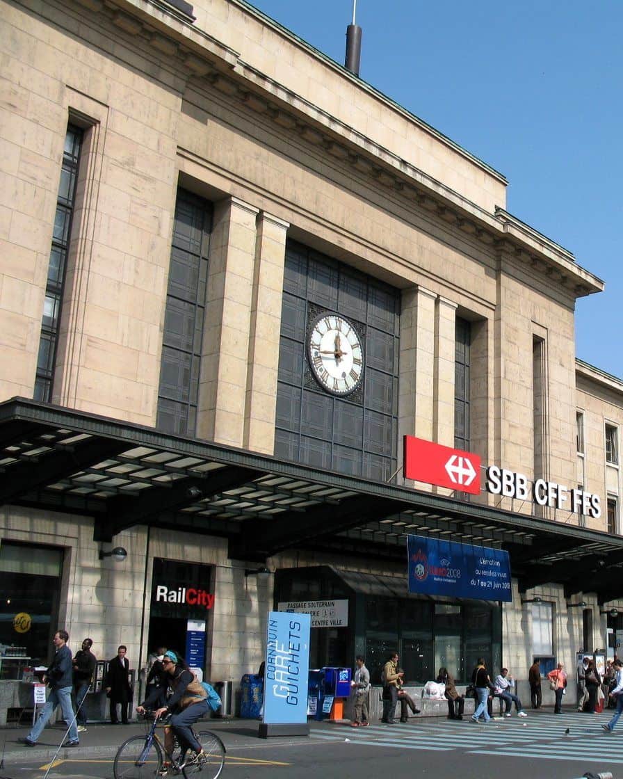 Façade de la gare de Genève-Cornavin, place de Cornavin, avec un tram et des voyageurs devant l'entrée principale